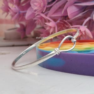 Puede incluir: Una pulsera de plata con un diseño de lazo único. La pulsera está hecha de metal brillante y presenta un detalle en forma de nudo en el lazo. El fondo incluye un objeto colorido con temática de arcoíris y flores rosas.