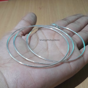 May include: Three silver bangle bracelets stacked on top of each other. The bracelets are thin and have a smooth, polished finish.