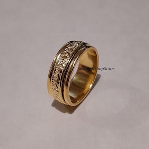 May include: A gold-colored ring with a textured band and a smooth, polished interior. The ring features an intricate floral pattern on the outer band. The ring is designed to spin. The ring is likely made of metal.