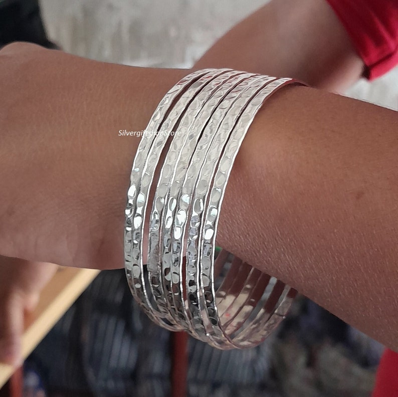 Pu&ograve; includere: Un set di sette braccialetti d'argento con una texture martellata. I braccialetti sono impilati sul polso di una persona.