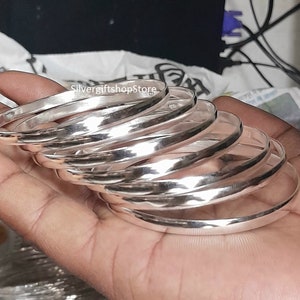 May include: A set of seven silver bangles stacked on top of each other. The bangles are smooth and shiny, and they have a slightly rounded shape.
