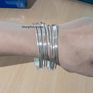 Op de afbeelding: Een set zilveren armbanden, gestapeld op de pols van een persoon. De armbanden zijn glad en glanzend, met een licht afgeronde vorm.