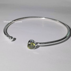 Puede incluir: Un brazalete de plata con una sola piedra preciosa verde engastada en el centro. El brazalete está abierto y tiene un extremo redondeado.