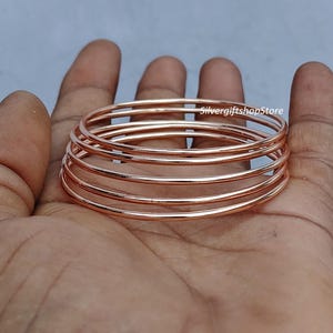May include: A stack of six thin, rose gold bangles. The smooth, metallic bracelets are stacked on top of each other, creating a layered effect. The image is taken from a close-up angle, highlighting the details of the jewelry. The text "SilvergiftshopStore" is visible.