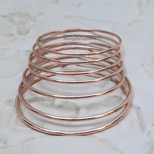 May include: Set of seven rose gold bangles stacked on top of each other. The bangles are thin and have a smooth, polished finish.