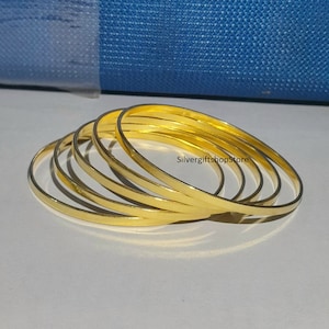 Puede incluir: Un conjunto de seis brazaletes de color dorado apilados uno encima del otro. Los brazaletes son lisos y brillantes.