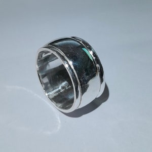 Puede incluir: Un anillo de plata con una banda ancha y un diseño texturizado. La banda está ligeramente curvada y tiene una línea verde sutil que recorre la parte superior.