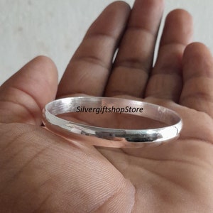 May include: A silver bangle bracelet with a smooth, polished finish. The bracelet is shown in a person's hand.