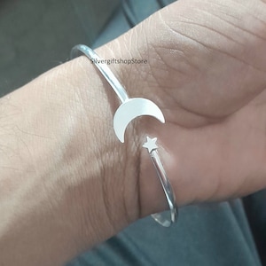 Può includere: Un braccialetto rigido in argento con un ciondolo a forma di luna crescente e stella. Il braccialetto è aperto e regolabile.