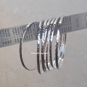 Pu&ograve; includere: Un set di otto braccialetti in argento martellato. I braccialetti sono impilati uno sopra l'altro e hanno tutti la stessa larghezza. I braccialetti sono in argento e hanno una finitura martellata. I braccialetti sono di dimensioni diverse, con il braccialetto pi&ugrave; piccolo che ha un diametro di 3,175 mm e il braccialetto pi&ugrave; grande che ha un diametro di 24,60625 mm.