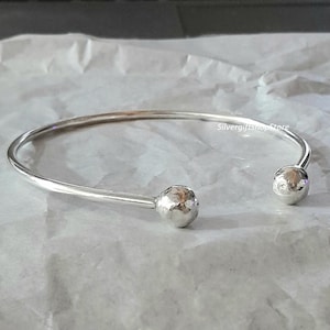 Könnte beinhalten: Ein silbernes Armreif-Armband mit gehämmerten Kugelenden. Das Armband ist offen und hat ein schlichtes Design.