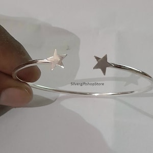 May include: A silver bangle bracelet with two star-shaped charms. The bracelet is open and has a simple design.