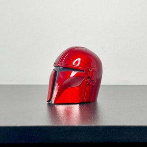 Puede incluir: Un casco mandaloriano rojo con detalles negros. El casco está hecho de un material brillante y liso.