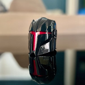 Puede incluir: Casco negro con una franja roja en la parte delantera. El casco tiene un acabado brillante y está detallado con líneas y ranuras intrincadas.