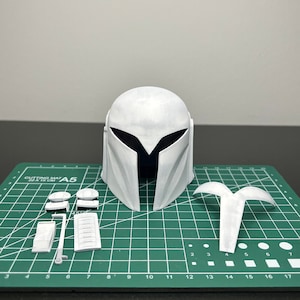 May include: A white Mandalorian helmet and its components are arranged on a green cutting mat. The helmet is the focal point, with its distinctive T-shaped visor. Additional parts include shoulder pads and other accessories.