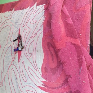 May include: A large pink fabric with a white design is being worked on with two sewing machines. The fabric is hanging on a wall and the machines are attached to the fabric with cords.