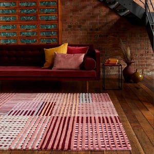 May include: A geometric patterned rug in shades of pink, red, and white dominates the foreground. A burgundy velvet sofa with colorful pillows sits against a brick wall. A small side table and decorative vases complete the room's decor.