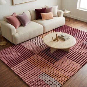 May include: A large area rug featuring a geometric grid pattern in shades of burgundy, pink, and orange. The rug is placed under a cream-colored sofa with several velvet throw pillows in coordinating colors and a round light-colored wooden coffee table.