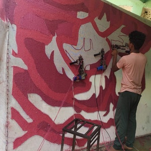 May include: A person uses a tool to create a large-scale abstract art piece on a wall. The art features a white background with red and pink abstract shapes.