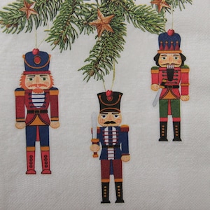 2 o 4 pezzi di tovaglioli di carta per lavori artigianali, tre statuette di giocattoli di schiaccianoci di Natale appese su rami di abete su sfondo beige con decorazioni a forma di stella