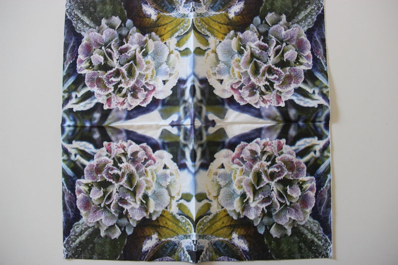 Puede incluir: Una servilleta cuadrada blanca con un dise&ntilde;o sim&eacute;trico de cuatro flores de hortensia. Las flores son una mezcla de morado, blanco y verde, y parecen estar cubiertas de escarcha. El fondo es azul oscuro y verde.
