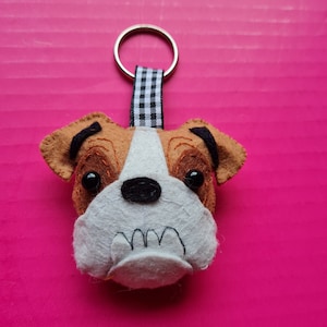 Könnte beinhalten: Ein handgefertigter Schlüsselanhänger in Form einer Bulldogge. Die Bulldogge ist aus braunem und weißem Filz gefertigt, mit schwarzen Augen und einer schwarzen Nase. Er hat einen silbernen Ring und ein schwarz-weiß kariertes Band.