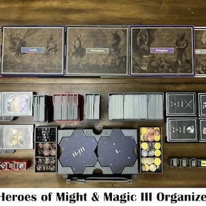 May include: A Heroes of Might & Magic III game organiser with multiple compartments for storing cards, tokens, and tiles. The organiser is made of black plastic and has a handle for easy carrying. The organiser is shown on a wooden table.
