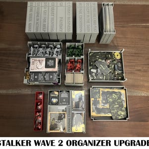 Puede incluir: Un organizador de juegos de plástico gris con múltiples compartimentos, que contiene piezas de juego, cartas y miniaturas. El organizador está etiquetado con títulos de juegos como "The Steel Giant" y "Personal Stories". El texto "STALKER WAVE 2 ORGANIZER UPGRADE" está en la parte inferior.