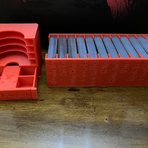 K&ouml;nnte beinhalten: Ein Set aus leuchtend orangefarbenen, 3D-gedruckten Kartenorganisatoren f&uuml;r ein Brettspiel. Ein Organizer hat mehrere F&auml;cher und eine Schublade mit der Aufschrift "XP" und "Saved Campaign". Der andere Organizer enth&auml;lt einen Stapel Karten.