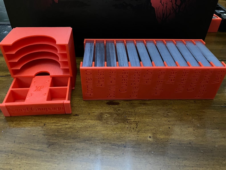 K&ouml;nnte beinhalten: Ein Set aus leuchtend orangefarbenen, 3D-gedruckten Kartenorganisatoren f&uuml;r ein Brettspiel. Ein Organizer hat mehrere F&auml;cher und eine Schublade mit der Aufschrift "XP" und "Saved Campaign". Der andere Organizer enth&auml;lt einen Stapel Karten.