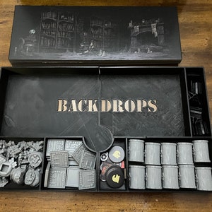 K&ouml;nnte beinhalten: Eine schwarze Spielbox mit dem Wort "BACKDROPS" in der Mitte. Die Box enth&auml;lt verschiedene graue Spielteile, darunter F&auml;sser, Bausteine und Spielsteine. Der Deckel zeigt eine detaillierte Illustration einer Bibliothek und eines Kamins.