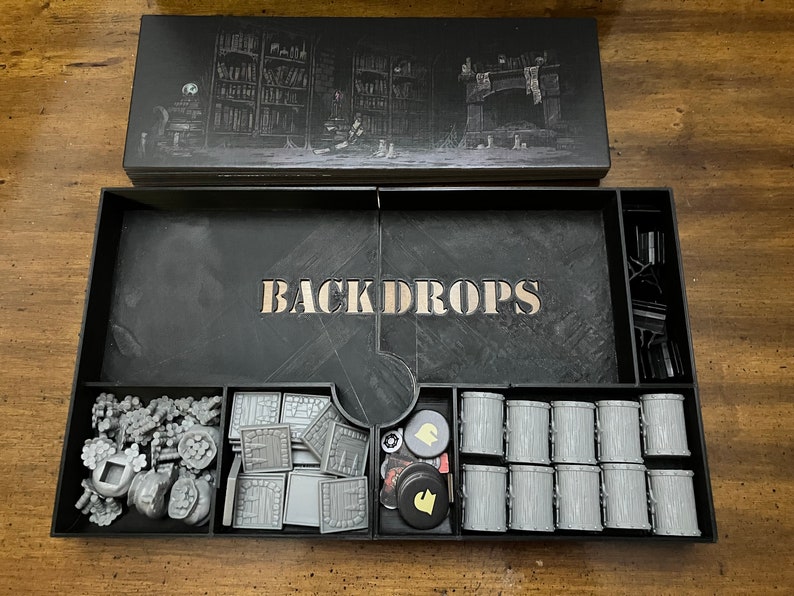 K&ouml;nnte beinhalten: Eine schwarze Spielbox mit dem Wort "BACKDROPS" in der Mitte. Die Box enth&auml;lt verschiedene graue Spielteile, darunter F&auml;sser, Bausteine und Spielsteine. Der Deckel zeigt eine detaillierte Illustration einer Bibliothek und eines Kamins.