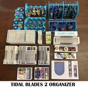 May include: A collection of board game organizers in white and light blue. The organizers are designed to hold cards, game pieces, and miniatures. The text "TIDAL BLADES 2 ORGANIZER" is printed at the bottom of the image.