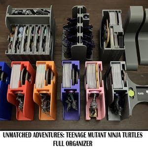 May include: A collection of gray, blue, orange, purple, and pink plastic game organizers. The organizers are designed to hold cards and game pieces. The text at the bottom reads "UNMATCHED ADVENTURES: TEENAGE MUTANT NINJA TURTLES FULL ORGANIZER."