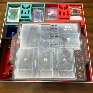 Op de afbeelding: Een witte, rode en blauwe spelorganizer met een transparante kunststof insert voor spelstukken. De insert heeft een rasterpatroon en de tekst "MAX 10" erop gedrukt.