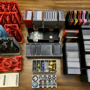 May include: A collection of plastic storage containers for board game components. The containers are in various colors, including red, blue, gray, and black. The containers are filled with miniature figures, cards, and tokens.