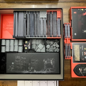 K&ouml;nnte beinhalten: Ein Brettspiel-Organizer mit orangefarbenen und grauen Komponenten. Der Organizer enth&auml;lt Kartenhalter, W&uuml;rfelschalen und F&auml;cher f&uuml;r Spielsteine. Die Spielbox ist ge&ouml;ffnet und zeigt den organisierten Inhalt, spielbereit.