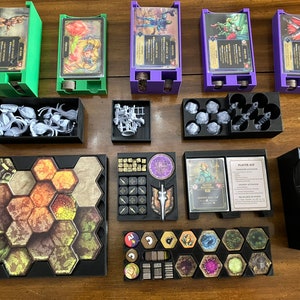 Puede incluir: Un conjunto de organizadores de plástico negro para componentes de juegos de mesa. Los organizadores incluyen compartimentos para cartas, dados, fichas y miniaturas. Los organizadores están dispuestos sobre una mesa de madera.