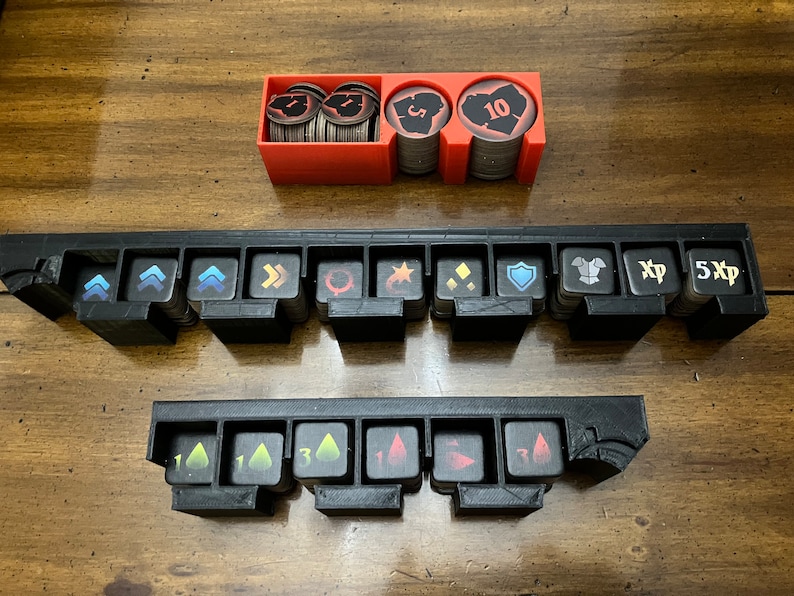 K&ouml;nnte beinhalten: Ein Set aus schwarzen und roten Spielorganisatoren f&uuml;r Brettspiel-Token. Die Organizer umfassen Schlitze f&uuml;r verschiedene Token mit Symbolen und Zahlen sowie einen roten Beh&auml;lter f&uuml;r M&uuml;nzen. Die Token haben Symbole wie Pfeile, Schilde und Erfahrungspunkte.