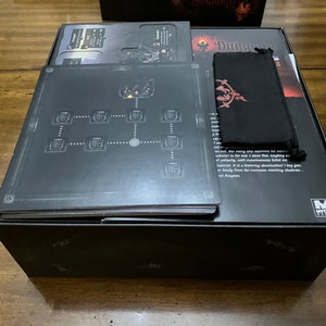 K&ouml;nnte beinhalten: Eine schwarze Brettspielbox mit verschiedenen Komponenten. Die Box enth&auml;lt Regelb&uuml;cher, Spielbretter und einen schwarzen Beutel mit rotem Emblem. Die Spielbretter zeigen ein Kartendesign.