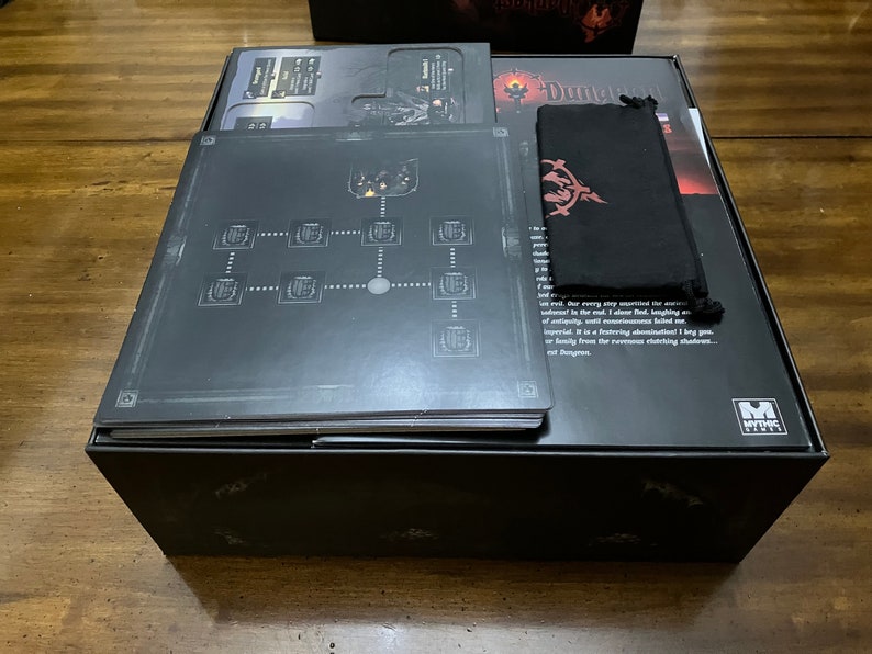 K&ouml;nnte beinhalten: Eine schwarze Brettspielbox mit verschiedenen Komponenten. Die Box enth&auml;lt Regelb&uuml;cher, Spielbretter und einen schwarzen Beutel mit rotem Emblem. Die Spielbretter zeigen ein Kartendesign.