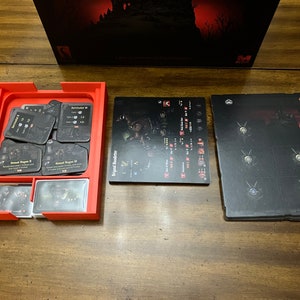 K&ouml;nnte beinhalten: Ein roter Spiel-Organizer mit verschiedenen Spielkarten und Spielbrettern. Die Karten sind schwarz mit wei&szlig;em Text und Grafiken. Die Spielbretter sind schwarz mit wei&szlig;en und roten Grafiken. Die Spielbox ist im Hintergrund sichtbar.