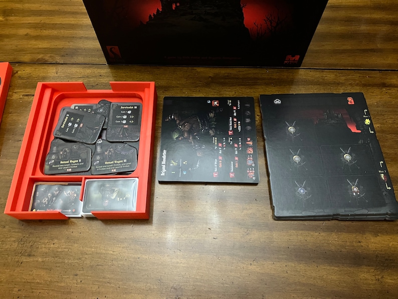 K&ouml;nnte beinhalten: Ein roter Spiel-Organizer mit verschiedenen Spielkarten und Spielbrettern. Die Karten sind schwarz mit wei&szlig;em Text und Grafiken. Die Spielbretter sind schwarz mit wei&szlig;en und roten Grafiken. Die Spielbox ist im Hintergrund sichtbar.