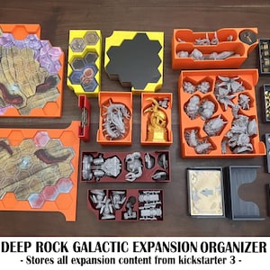 May include: A board game organizer set for Deep Rock Galactic Expansion, featuring orange, yellow, red, and grey plastic trays. The set includes hexagonal tile holders, miniature storage bins, and card organizers with the text DEEP ROCK GALACTIC EXPANSION ORGANIZER - Stores all expansion content from kickstarter 3 -.