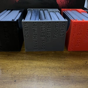 K&ouml;nnte beinhalten: Drei 3D-gedruckte Kartenorganisatoren in Schwarz, Grau und Rot, gef&uuml;llt mit Karten. Jeder Organizer ist mit spielbezogenen Begriffen wie "Quests", "Monster" und "R&auml;ume" beschriftet. Die Organizer dienen zum Sortieren und Aufbewahren von Spielkarten.