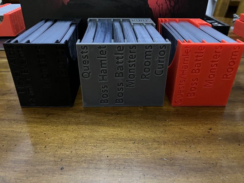 K&ouml;nnte beinhalten: Drei 3D-gedruckte Kartenorganisatoren in Schwarz, Grau und Rot, gef&uuml;llt mit Karten. Jeder Organizer ist mit spielbezogenen Begriffen wie "Quests", "Monster" und "R&auml;ume" beschriftet. Die Organizer dienen zum Sortieren und Aufbewahren von Spielkarten.