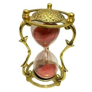 Puede incluir: Un reloj de arena dorado con una base adornada y un bulbo de vidrio transparente lleno de arena roja. El reloj de arena es un objeto decorativo.