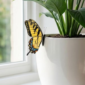 May include: A white ceramic plant pot with a green plant and a yellow butterfly with black stripes and blue details. The butterfly rests on the pot's edge. The pot sits on a white windowsill next to a window.