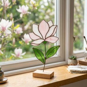 Pode incluir: Uma escultura de flores em vitral com uma flor rosa e branca, folhas verdes e uma base de madeira. A flor é exibida em uma janela com um fundo desfocado de flores rosas e uma janela.