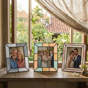 K&ouml;nnte beinhalten: Drei dekorative Fotorahmen mit R&auml;ndern im Glasmalerei-Stil. Die Rahmen sind in Blau-, Pastellgelb- und Rosat&ouml;nen gehalten und halten jeweils ein Foto. Die Rahmen werden auf einer Holzoberfl&auml;che in der N&auml;he eines Fensters ausgestellt.